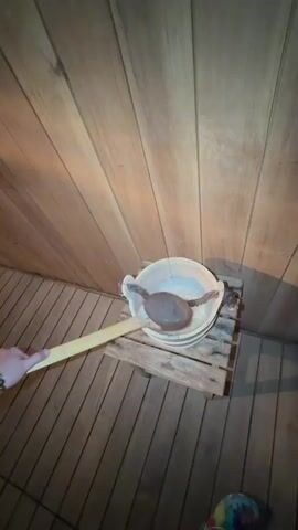 A person's hand, holding a wooden ladle filled with water, is seen in the foreground of a sauna. The ladle is being used to pour water onto a pile of hot stones. The sauna has wooden walls and a metal stove. The water sizzles and creates steam as it hits the hot stones. The video shows a close-up view of the ladle and the stones, and then pans out to show more of the sauna environment.