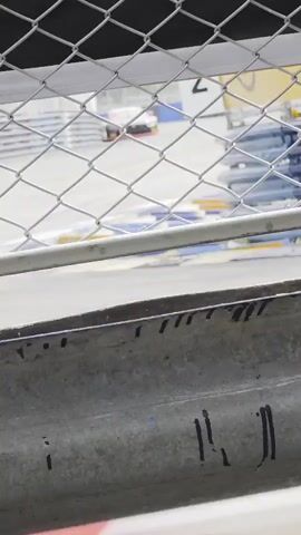 A race car speeds past on a track, viewed through a chain-link fence. The car is white with red and black accents. The track has yellow and blue markings. The foreground shows a concrete barrier with dark streaks.