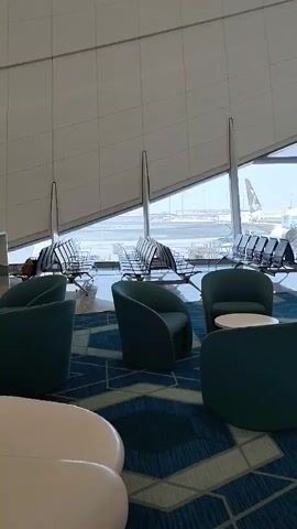 The video shows an airport terminal waiting area. In the foreground, there are several plush, teal-colored armchairs arranged around small, round white tables. A large, white, organically shaped bench or seating area is partially visible in the bottom left corner. The floor is covered in a blue carpet with a geometric pattern. In the mid-ground, there are rows of standard airport seating with teal-colored cushions, facing large windows that look out onto an airport tarmac. An airplane is visible through the windows. Beyond the seating, there are tall, silver cylindrical trash receptacles and some metallic pillar structures. The walls of the terminal are a light color, and the ceiling is angled and white. The lighting appears to be natural daylight coming through the windows. There are no people visible in the video.