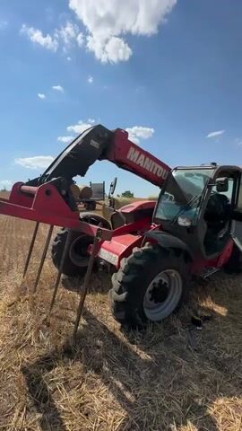 The video shows a Manitou telehandler tractor with its front right door open and a flat tire. Tools, including a wrench and a drill, are laid out on the ground near the flat tire, suggesting that someone is in the process of changing it. The tractor is parked in a dry, grassy field with hay bales visible in the background. The sky is blue with some scattered clouds. The camera pans across the scene, focusing on the damaged tire and the tools.