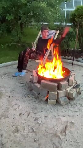A person with short, dark hair and a black shirt sits in a striped lounge chair next to a fire pit. The fire pit is made of bricks and contains a blazing fire. The person is holding a bottle and appears to be looking at the camera. The scene is outdoors, with trees, bushes, and a greenhouse in the background. The ground is covered in sand and gravel.