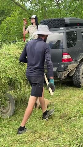 In Navdaraantkari, Georgia, a grassy outdoor scene unfolds with a black SUV parked amidst lush greenery. A woman in a camouflage t-shirt and jeans stands near the rear of the SUV, holding a smartphone. A man, wearing a navy long-sleeved shirt, black shorts, and a light-colored hat, is actively engaged in raking hay. He is wearing blue gloves and athletic shoes. A small puppy, light brown in color, is present in the scene, navigating through the grass. The SUV is equipped with a roof rack featuring a spare tire and a storage box. The background consists of dense trees, suggesting a rural or wooded environment. The lighting indicates it is daytime, and the presence of cut grass suggests haymaking or similar agricultural activity is underway. No visible text is present on the people or the dog. The license plate of the SUV is visible and reads "I 022 YY".