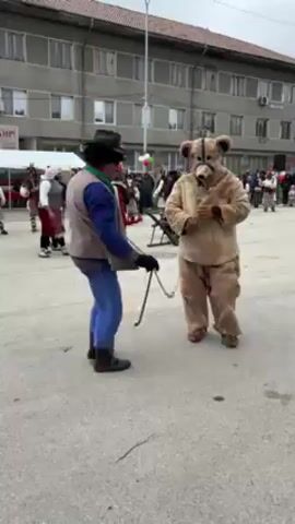 Here's a description of the video:

The video captures a street celebration. A person in a bear costume is led on a chain by another person dressed in a blue shirt, vest, and black hat. They stand in a town square or street, with a group of people in the background. The background includes a building, tents, and decorations with the Bulgarian flag. There's an event taking place with music playing. People in traditional costumes can be seen in the background dancing.