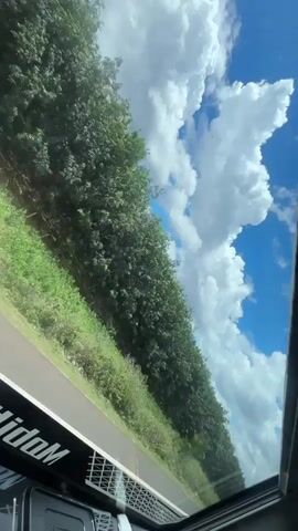 The video shows a view from inside a vehicle driving alongside a road. The camera is angled upward, capturing a line of dense trees alongside the road, a bright blue sky, and scattered, fluffy white clouds. The road has a white line marking the edge of the lane. The vehicle appears to have a glass roof or sunroof. Part of the vehicle's interior is visible at the bottom of the frame, including a section with the word "MohidoM" printed on it. As the vehicle moves, a truck is seen passing by, partially obscuring the view with its black trailer marked with a white and blue graphic design and some writing in what appears to be Vietnamese. The primary focus remains on the outdoor scenery through the roof of the car.