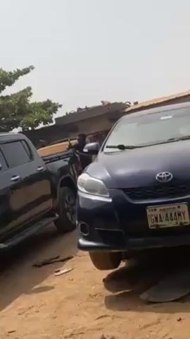 The scene shows a dirt lot with three vehicles parked. In the foreground is a dark blue Toyota Matrix with a license plate reading "GWA-444MY." Next to it, to the left, is a black pickup truck. A person is standing near the rear of the pickup truck, wearing a dark shirt. In the background, there's a building and some trees. The weather appears sunny. A sound like a hammer hitting metal can be heard.