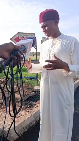 A man wearing a white traditional outfit and a maroon kufi hat is being interviewed outdoors. He is standing on a dirt path with a concrete border. Behind him, there is a sign pointing to the right, which appears to read "AMBASSADOR UBA UNIVERSITY, GAMA NAERLS SUITES & CONFERENCE." A hand is holding a microphone with a fluffy windscreen towards the man. The background shows a grassy area and a hazy sky. The man is gesturing with his hands as he speaks.