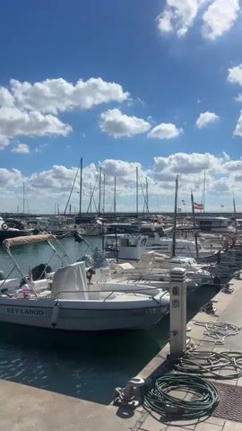 The video is a vertical shot of a marina on a sunny day with a blue sky and scattered white clouds. The foreground features a concrete pier with coiled ropes and a bollard. In the water, several boats are docked. The closest boat is a white motorboat with the text "KEY LARGO" visible on its side. Behind it are more motorboats and sailboats, with numerous masts extending upwards. In the background, the horizon is visible, with more boats and possibly land in the distance. Flags are also visible on some of the boats. The overall scene depicts a tranquil harbor environment.