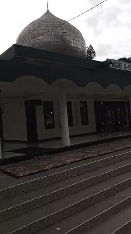 The media file shows a view of Masjid Al-Munawwarah in Blang Bebangka, Indonesia. The mosque features a large, silver dome and a covered walkway supported by pillars. The building is situated on a hillside with lush green trees and vegetation visible in the background. In front of the mosque, there is a wide set of stairs leading down to a river. The river is flowing with visible ripples and waves, suggesting a moderate current.

A few people are visible in the scene. One person is standing on the steps, appearing to look out at the river or the surrounding landscape. Another person is near the entrance of the mosque. The overall atmosphere appears calm and serene.

The sky is overcast, indicating a cloudy or possibly rainy day. The time of day is difficult to determine precisely but appears to be daytime. The combination of the mosque, the river, and the natural landscape suggests a peaceful and spiritual setting. The visible text on the mosque reads "MASJID AL-MUNAWWARAH". The presence of the river and the surrounding greenery hints at a location with natural beauty.