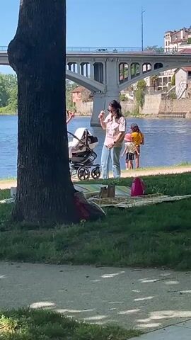 The video shows a public park on a sunny day, with a bridge visible in the background over a body of water. A large tree trunk dominates the left side of the frame. People are moving about near the water's edge. Two women are interacting near a stroller, one wearing white pants and the other wearing blue jeans. A couple, one in a green shirt and the other in white shorts, walks past them. There is a young child in a yellow shirt standing by the water. A picnic blanket and assorted items are scattered on the grass near the tree.