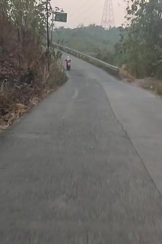 A person is riding a motorcycle on a paved road. The road curves to the left and goes uphill. There are trees and vegetation on the left side of the road, and a guardrail on the right side. In the background, there are hills and a large electrical tower. The sky is overcast. The time is dawn or dusk. The motorcycle is red and appears to be moving at a moderate speed.