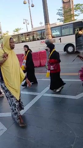 The video shows a busy outdoor pedestrian area. A group of women in religious attire walks along the sidewalk. They are dressed in black abayas and hijabs, with yellow scarves around their necks indicating they are with "ASIA TOUR." Some carry pink bags. A city worker in a blue uniform sweeps the sidewalk with a broom. A large white bus with a prominent "1" on the side is parked in the background. A clock tower is also visible. The day appears bright and sunny. There are other pedestrians in the background.