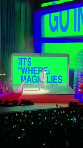A female performer stands center stage on a raised platform, dressed in a white dress and white knee-high socks. She is holding a microphone and singing. Behind her is a large screen shaped like an open laptop, displaying the words "GO INSIDE IT'S WHERE THE MAGIC LIES" in bright green and blue letters. To the left of the platform is a large, red, banana-shaped inflatable object. To the right, two figures in orange outfits are dancing. The background features a large screen with bright blue and green lights and horizontal lines. The foreground shows a dark crowd of people holding up cell phones. The scene appears to be a live concert performance.