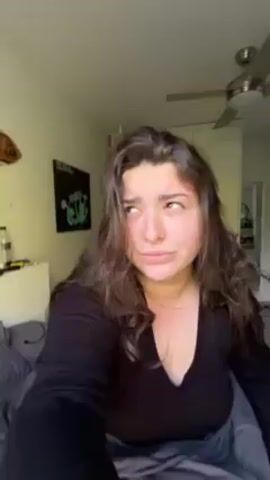 A woman with long, wavy brown hair and a black shirt is in a bedroom. The room has light yellow walls, a ceiling fan, a bed with gray bedding, a white nightstand with items on it, a wardrobe, and a poster on the wall. The woman looks at the camera with a somewhat sad expression. The camera moves to show a window with green bars. Outside the window are green trees and a blue sky with some clouds.