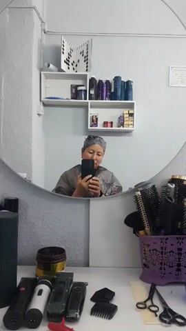 A person is reflected in a mirror in a room that appears to be a hair salon. The person, wearing a grey smock, is holding a smartphone and taking a selfie. Their hair is wrapped in a grey cap. On shelves behind the person are various bottles and containers, likely hair products. In the foreground, on a white surface, are several hair styling tools, including clippers, scissors, brushes in a purple basket, and spray bottles. The background wall is painted white with some textured grey paint.