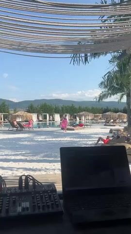 The video shows a view from behind a DJ setup, which includes a laptop and a mixer board, looking out onto a crowded beach resort. Several cabanas with thatched roofs and white curtains are visible, along with lounge chairs occupied by people sunbathing. A swimming pool is also present, with some individuals in the water. The beach area is covered in white sand, and there are palm trees and lush green mountains in the background under a clear blue sky. The overall atmosphere suggests a lively and relaxed daytime event.