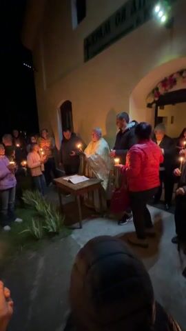 Sure, here is a detailed description of the video:

The video shows an outdoor religious ceremony at night, presumably Eastern Orthodox, due to the vestments and style of the priest. The priest, wearing a white and gold robe and a large cross, holds a lit candle bundle and speaks from an open book on a small wooden table. Several people stand around him, most holding lit candles. The setting is against a light-colored building wall. The ambiance is subdued, focusing on the priest’s actions and speech.