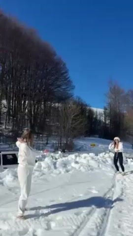Two women are playing catch with snowballs in a snowy, outdoor scene on a bright, sunny day. The woman on the left is wearing a white sweatsuit, while the woman on the right is wearing a white jacket, black pants, and a white hat. They are standing on a snow-covered path with trees on either side and a hill in the background. A silver car is partially visible behind a snowbank. The woman in white misses a snowball and the woman recording asks if she recorded it.