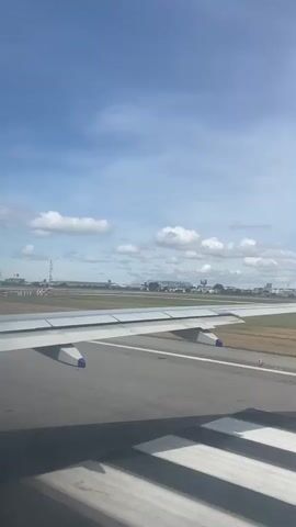 The video shows a view from inside an airplane, looking out the window at the wing and the runway. The plane is moving, presumably during takeoff or landing. The sky is blue with scattered white clouds. There are buildings and structures visible in the distance, suggesting an airport or urban area. The runway is marked with white lines.