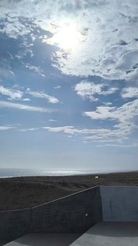 The video shows a coastal scene with a gray gravel area in the foreground, followed by a tan, desolate landscape, and a mountain range in the distance, under a cloudy sky. Initially, the ocean is visible in the distance but becomes obscured as the camera pans to the right, focusing on the gravel area and the distant mountain range. The scene is mostly devoid of color, consisting primarily of gray, tan, and muted blue hues. There are no people or animals visible in the frame. The camera seems to be stationary, capturing the scene from an elevated position, possibly from a structure overlooking the landscape.