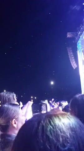 The video shows a crowd of people at a night event, possibly a concert or festival. The sky is dark, with some lights visible in the distance, possibly stage lights or streetlights. Many people are holding up their phones, likely to take pictures or videos of the event. The crowd is dense, with heads and shoulders filling the frame. The overall impression is one of excitement and engagement with the event. The audio features music and crowd noise.