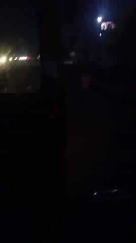 This is a shaky, dark video likely filmed at night. The camera appears to be in motion, possibly in a vehicle. There are streetlights visible, casting a harsh glow on what looks like a road or dirt path. In the background, there are dark shapes that could be trees or buildings. The overall impression is of a dimly lit, possibly rural or suburban environment at night. There are no clear people or specific objects that can be identified due to the poor lighting and motion blur.