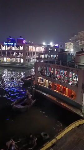 The video shows a nighttime harbor scene. Several multi-story boats or ferries are docked, illuminated by lights. People are visible on the boats, some walking and some gathered in groups. A small boat is near one of the ferries. The water reflects the lights. In the background are buildings on a hillside. The sky is dark.