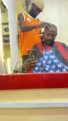 A barber is giving a haircut to a customer in a barbershop. The barber, wearing a tan beanie and an orange jersey, is using clippers on the customer's head. The customer, who has a beard, is sitting in a barber's chair draped with a cape featuring the American flag. He is holding a smartphone and appears to be taking a selfie. In the background, there is a mirror, a fan, and a shelf with electronic equipment. The room is brightly lit.