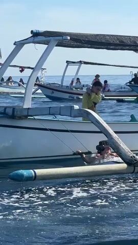The video shows two women and other people on boats in a body of water under a sunny sky. A woman in a yellow cover-up stands on the front edge of a white boat, while another woman in a pink top holds onto the floatation device on the side of the boat and bobs up and down in the water.  Several similar boats are visible in the background, some with passengers. The scene appears to be set in a touristy area with boats used for recreational water activities.