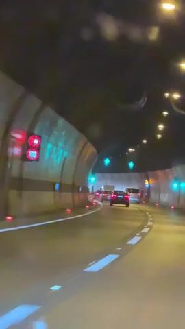 The video shows a first-person perspective of driving through a tunnel at night. The tunnel walls are illuminated with green and blue lights, and overhead lights cast a warm glow on the asphalt road. White lane markings guide the way, and red markers are visible along the sides of the road. Several cars are driving ahead, their taillights visible in the distance. The tunnel appears to be curving to the right.