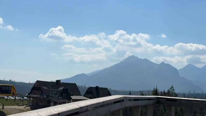 The media file captures a panoramic view of a mountainous landscape under a clear blue sky dotted with fluffy white clouds. The scene is set on a wooden platform, indicated by the wooden railing in the foreground, which suggests an elevated vantage point, possibly a walkway or viewing deck.

In the middle ground, several traditional-style buildings with dark roofs and wooden accents are visible. One prominent building appears to be a chalet or guesthouse, with visible balconies and architectural details. To the left of these buildings, a bright yellow object, likely part of a cable car or ski lift station, is partially visible.

The background is dominated by a majestic mountain range that stretches across the horizon. The mountains are layered, with the closest peaks appearing a deeper blue-grey, and successive ranges fading into lighter shades due to atmospheric perspective. Some snow patches can be discerned on the higher peaks, even in what appears to be a warmer season given the lush green trees in the lower elevations. A dense forest of evergreen trees carpets the foothills of the mountains.

The time of day appears to be late morning or early afternoon, with bright sunlight casting shadows and illuminating the scene. The weather is fair and sunny, with a pleasant, clear sky. The overall mood is serene and majestic, showcasing the natural beauty of the mountainous region. The location is identified as Małe Ciche, Poland, a place known for its natural scenery and outdoor activities in the Tatra Mountains. There is no visible text within the media file.