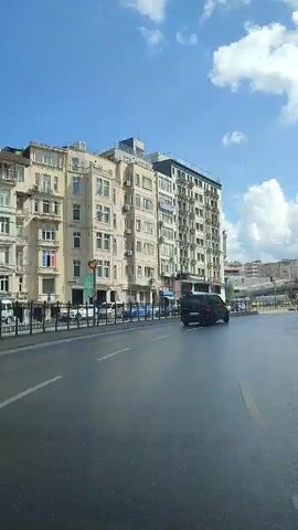 A video captures a street scene with buildings on the left and right sides. On the left, there are multi-story buildings with many windows and balconies. Several cars, including a dark van and a blue car, are visible on the road. The road appears wet, suggesting recent rain or cleaning. On the right side of the road, more buildings are visible, some under construction with scaffolding. Several yellow taxis are also present on the road, and there are orange traffic cones in the foreground. The sky is blue with scattered white clouds. The video is shot from a moving vehicle, providing a dynamic perspective of the urban environment.