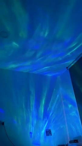 This video shows a corner of a room illuminated by a blue and green aurora borealis-like projector light. The light patterns are cast on the walls and ceiling, creating a dynamic and immersive effect. The room appears to be dimly lit, with the projector light being the primary source of illumination. There is a small framed picture hanging on the wall and what appears to be some electronic equipment in the bottom right corner of the frame. The video is shot from a low angle, looking up towards the ceiling.