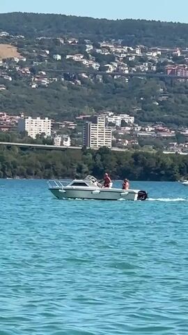 A white motorboat with two people on board sails across the turquoise water of a bay. The boat is moving from left to right. The person in the front appears to be steering, while the person behind them is seated. In the background, a lush green hillside is dotted with buildings and houses, indicating a coastal town or city. A bridge can be seen in the mid-ground, connecting two parts of the land. The sky is clear and blue, suggesting a sunny day.