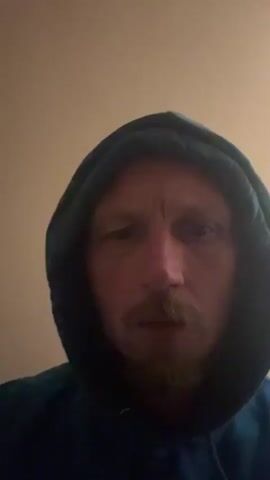 The video shows a close-up of a man's face, wearing a dark blue hooded sweatshirt. The man has light skin, a mustache, and a beard. His eyes are looking directly at the camera, and he appears to be speaking. The background is a plain, light-colored wall. The lighting is dim, casting shadows on the man's face.
