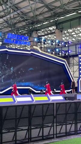 The video shows a stage set up indoors under a large metal roof with exposed beams and lighting rigs. A large LED screen behind the stage displays a dynamic blue watery graphic. In front of the screen, three performers in red flowing robes and masks are dancing. They are positioned on a raised platform in front of the main screen. To the right of the stage, a woman in black is standing and watching. The stage is surrounded by black metal barricades, and a green grassy area is visible in the foreground. The overall environment appears to be an exhibition hall or a large indoor venue prepared for a performance or event.