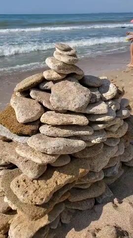 A low-angle medium shot shows a stack of flat, weathered stones carefully balanced on a sandy beach. The stones are light brown and tan, with some appearing to have a porous texture. The stack is conical in shape, wider at the base and tapering to a point at the top. In the background, the ocean waves gently roll onto the shore. A young child wearing a red hat and yellow shorts is visible on the beach to the right of the stone stack, appearing to play near the water's edge. The sky is a clear blue.
