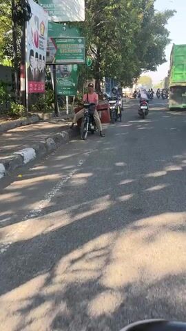 The video shows a street scene, likely in a city, with several motorbikes and a truck. In the foreground, a person is sitting on a motorbike, with their left leg on the ground. They are wearing a pink t-shirt and dark pants. The motorbike is black and appears to be a scooter-style. Behind this motorbike, another person on a motorbike, wearing a blue and white patterned shirt and a blue helmet, is looking at their phone. Further down the street, more motorbikes are visible, as well as a large green truck and a red truck. The environment appears to be a sunny day, with trees lining the street and some buildings in the background. The road is paved and there are white lines marking the lanes. The overall impression is of a busy street with moderate traffic.
