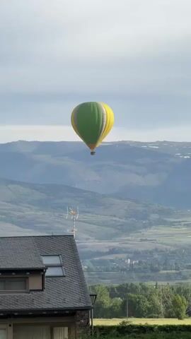 A green and yellow striped hot air balloon floats left to right across the sky, high above a distant mountainous and hilly landscape. In the foreground, the dark grey tiled roof of a building with a skylight and a small antenna array is visible on the left, partially obscured by a green hedge across the bottom of the frame. The sky is bright with some clouds.