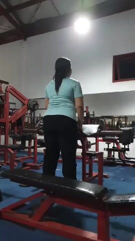 A woman in a light blue t-shirt and black pants is performing a barbell row exercise in a dimly lit gym. She is standing with her back to the camera, holding a barbell with both hands. The gym has red weight machines and blue mats on the floor. A bright light is shining from the ceiling.