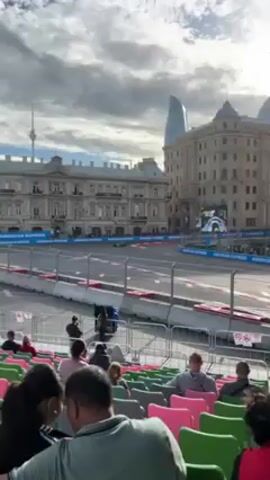 The video shows a Formula 1 race from a spectator's viewpoint in the stands. A race car speeds around a corner on the track, with buildings and the Baku Flame Towers visible in the background. The stands are filled with people watching the race.  "American Express" is visibly printed around the side of the race track. The weather appears cloudy with some sunlight peeking through.