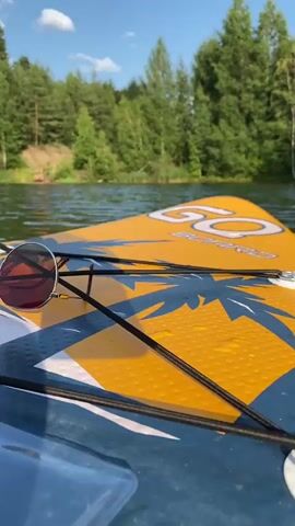 The video shows a close-up, first-person view from a paddleboard on a lake. The paddleboard is yellow with a blue and white palm tree design. A pair of sunglasses rests on the paddleboard. In the background, there is a lush green forest lining the lake and a bright blue sky with scattered clouds. The water is calm with gentle ripples. There is one person visible in the distance on the shore, near the trees. The camera pans slightly to show more of the lake and surrounding scenery.