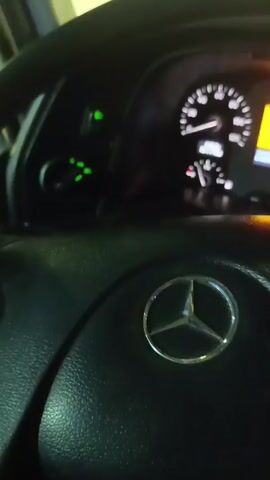 This media file provides a close-up view of a vehicle's interior, specifically focusing on the steering wheel and dashboard. The steering wheel prominently displays the Mercedes-Benz logo, suggesting it is a Mercedes-Benz vehicle, likely a truck given the dashboard design. The dashboard features a digital display and analog gauges, with various indicator lights illuminated, including a red hazard light and green turn signals. The time of day appears to be nighttime or indoors, as the interior lights are on and the dashboard displays are illuminated. The scene is focused on the driver's perspective, giving a sense of being inside the vehicle. There is no visible text that can be deciphered in the image. The context provided indicates the location as Қопа ауылдық округі, Kazakhstan. No people are visible, and the activity taking place is the vehicle's ignition or operation, indicated by the illuminated dashboard. The weather is not discernible.