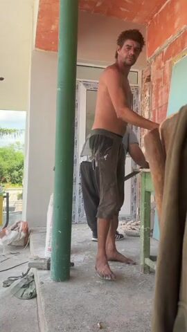 Two men are working outside a building. One man, shirtless, with a beard and dark hair, stands in the foreground, facing the camera. He wears dark pants, some of which are stained with paint or plaster. Another man, wearing a white cap, a gray t-shirt, dark pants, and a back brace, is seen from the back, working on a door frame in the middle ground. He holds a tool in his right hand. The building wall behind him is red brick and light blue drywall. A green pole is between the two men. In the background, there are trees and a sky. The ground is concrete, covered in dust and debris.