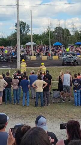 The video shows an outdoor demolition derby event. In the foreground, spectators are watching the event. In the middle ground, there are several destroyed vehicles, some of which are emitting smoke. Several individuals in safety vests and helmets, likely safety personnel or organizers, are present near the vehicles. In the background, there is a large crowd of spectators seated in bleachers, with tents and food vendors visible. The sky is cloudy. The overall atmosphere appears to be that of a lively event.