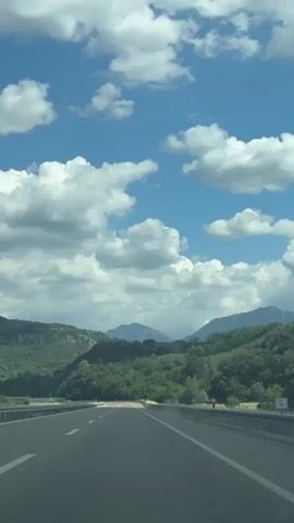 The video shows a view from a vehicle traveling on a highway. The road is paved and has white lane markings. On either side of the road, there are green, tree-covered hills and mountains. The sky is blue with fluffy white clouds. The overall impression is one of a scenic drive through a natural landscape.