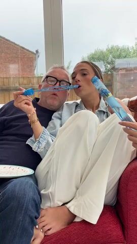 A man in glasses wearing a navy blue shirt and blue jeans sits on a red couch on the left. A young woman with long brown hair wearing a light blue denim shirt and white wide-leg pants sits next to him. They are holding two different types of bubble wands, one blue and one clear with a blue top. They are making bubbles and trying to touch them together. The man has a white plate with red dots on his lap. The background shows a large window with a brick house and green bushes outside.
