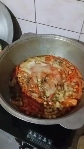 A close-up, high-angle shot shows a large, round, metal cooking pot situated on a black stovetop. The pot is filled with food, primarily consisting of shredded carrots, chickpeas, and what appears to be meat, all simmering in a liquid. The food is arranged in layers, with the carrots forming an outer ring and the chickpeas at the bottom. The surface of the food is slightly bubbly, suggesting it is actively cooking. The background is out of focus, but hints of a tiled wall can be seen. The lighting is somewhat dim, creating a warm, domestic atmosphere. The overall impression is that of a hearty meal being prepared.