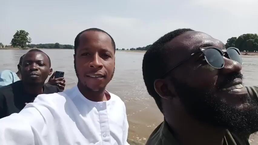 The video shows four men on a boat on a body of water, possibly a river or lake. The man in the white shirt appears to be holding the camera, taking a selfie. The man to his right is wearing sunglasses and a green shirt and is smiling. Another man in a black shirt with a colorful hat stands to the left of the man in the white shirt. A fourth man can be seen partially behind the man in the white shirt. The water is brown and appears shallow, with a sandy shoreline visible in the background, dotted with trees. The man in the green shirt is later seen holding a phone, possibly taking photos of the scenery. The overall atmosphere seems cheerful and relaxed.
