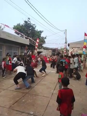 The video shows a group of people participating in a game of tug-of-war. The game is taking place in a street or alleyway, lined with buildings on either side. Red and white Indonesian flags are strung across the street, indicating a celebration or national holiday. A crowd of spectators, including adults and children, are gathered on both sides of the playing area, watching the event. The participants in the tug-of-war are evenly divided into two teams, with several people on each side pulling on a thick rope. The ground is paved with concrete tiles. The sky is overcast.