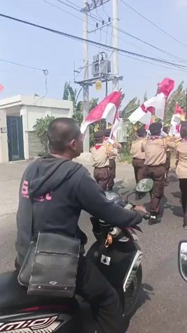 A group of scouts in uniform march in a parade, holding Indonesian flags. The scouts are wearing tan shirts, brown pants, and dark hats. They are carrying poles with red and white flags, which are fluttering in the wind. The parade is taking place on a paved road, with a building and trees visible in the background. The sky is clear and sunny. The scouts appear to be in formation and marching in an organized manner. Some of the scouts are wearing dark boots, while others are wearing dark shoes. There are power lines and a utility pole visible above the parade. The flags are held high and are a prominent feature of the scene. The camera is positioned at a low angle, looking up at the scouts. The video is in color and appears to be shot during the daytime.